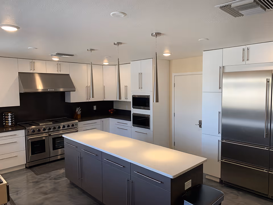 Modern kitchen with white upper cabinets and dark lower cabinets on the island. Stainless steel appliances including a large refrigerator, double oven, and stove with a vent hood. Three pendant lights hang above the island, and there is a door in the background.