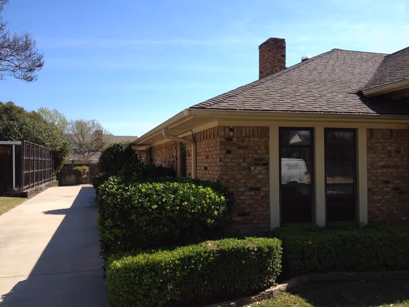 Exterior view of a single-story brick building with a sloped roof and chimney, surrounded by neatly trimmed bushes and a concrete walkway on the left side under a clear blue sky.