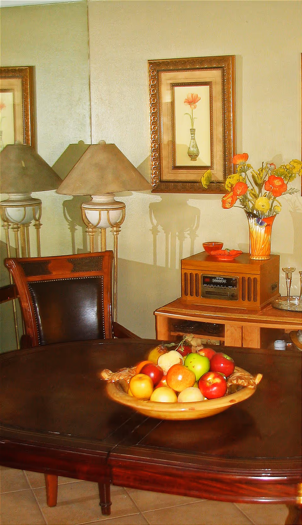 Dining room table with a bowl of fruit, a chair, lamps, framed floral artwork and a sideboard with a vase of flowers.