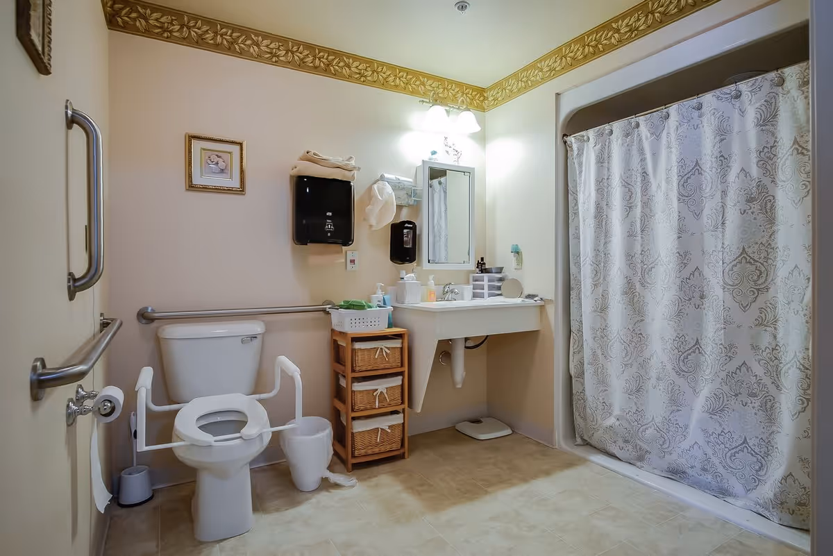 A clean and accessible bathroom with a toilet equipped with safety rails, a white sink with a mirror above it, a shower with a patterned curtain, a small wooden storage unit with baskets, and grab bars on the walls. The walls are light-colored with a decorative border near the ceiling.