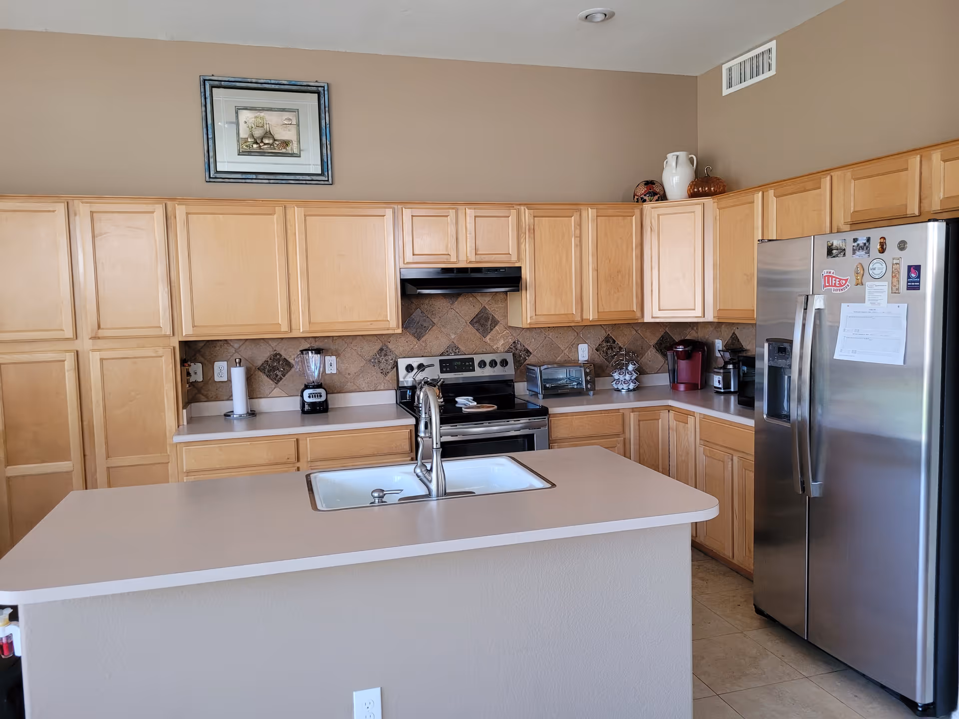 Bright kitchen with an island and sink, light wood cabinets, stainless steel refrigerator and stove, and a tiled backsplash.
