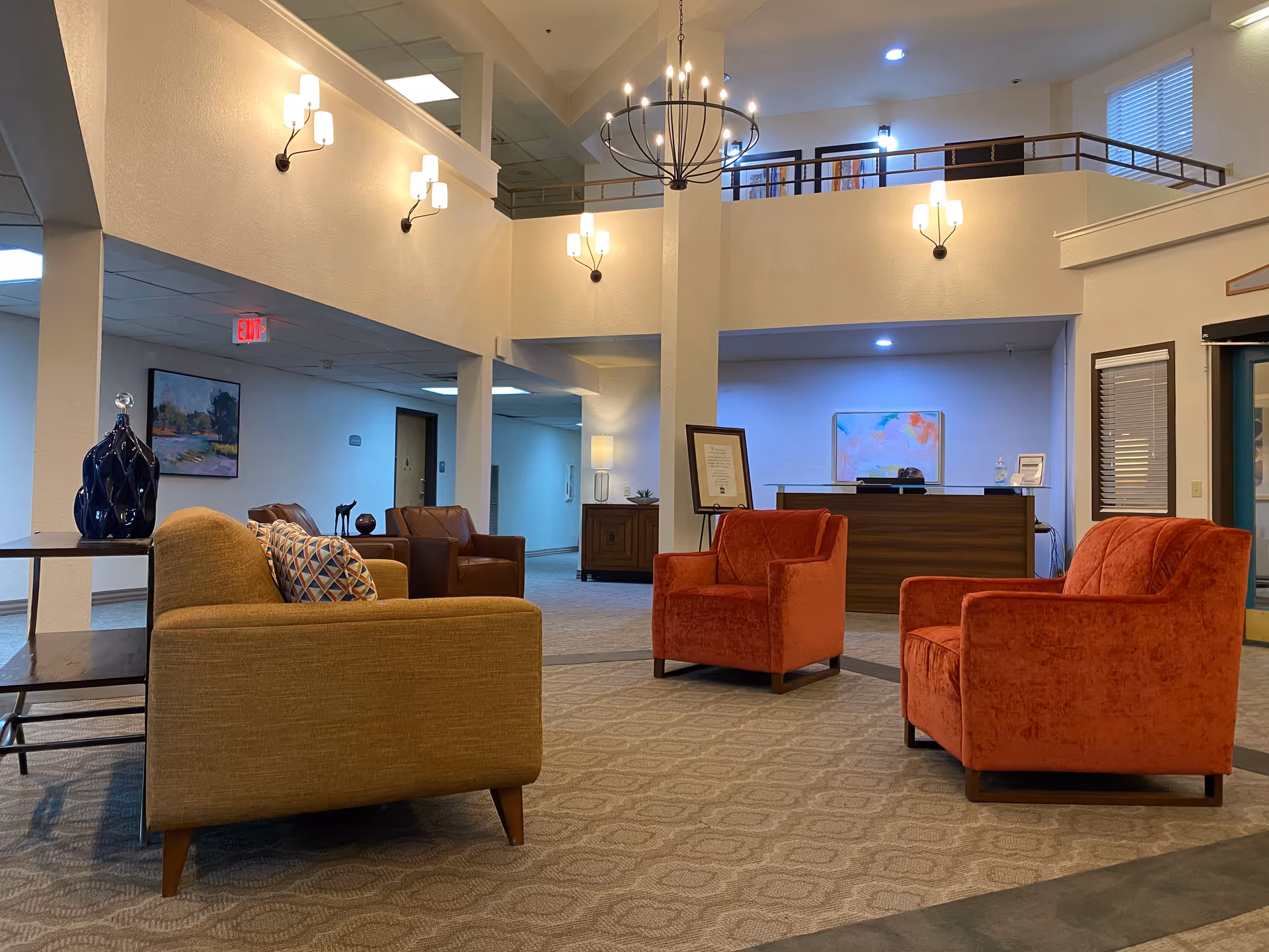 A spacious senior living facility lobby with a high ceiling and modern lighting fixtures. The seating area includes a mustard yellow sofa with a patterned cushion, two orange armchairs, and two brown leather chairs. A wooden reception desk is visible in the background with a colorful abstract painting on the wall behind it. The area is carpeted and decorated with a blue vase on a side table and framed artwork on the walls.