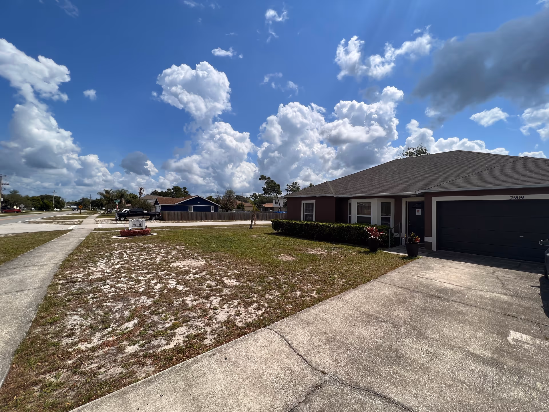 Single-story residential building with a dark roof and garage, surrounded by a patchy lawn and a concrete driveway under a partly cloudy blue sky.