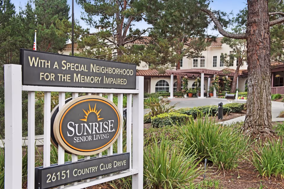A Sunrise Senior Living entrance sign in front of the landscaped driveway and facility building.