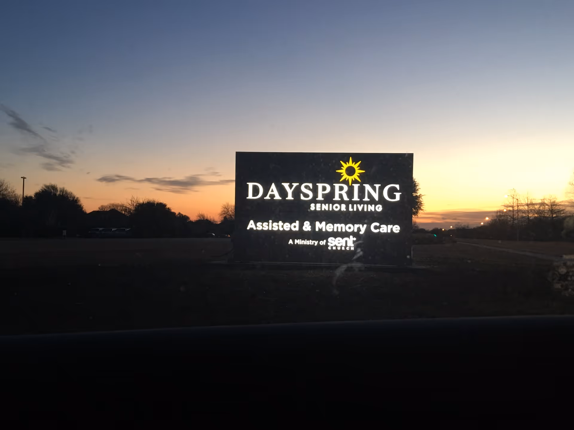 Illuminated sign for DaySpring Senior Living at dusk, displaying the text 'Assisted & Memory Care' and 'A Ministry of Sent Church' with a sun logo above the name.