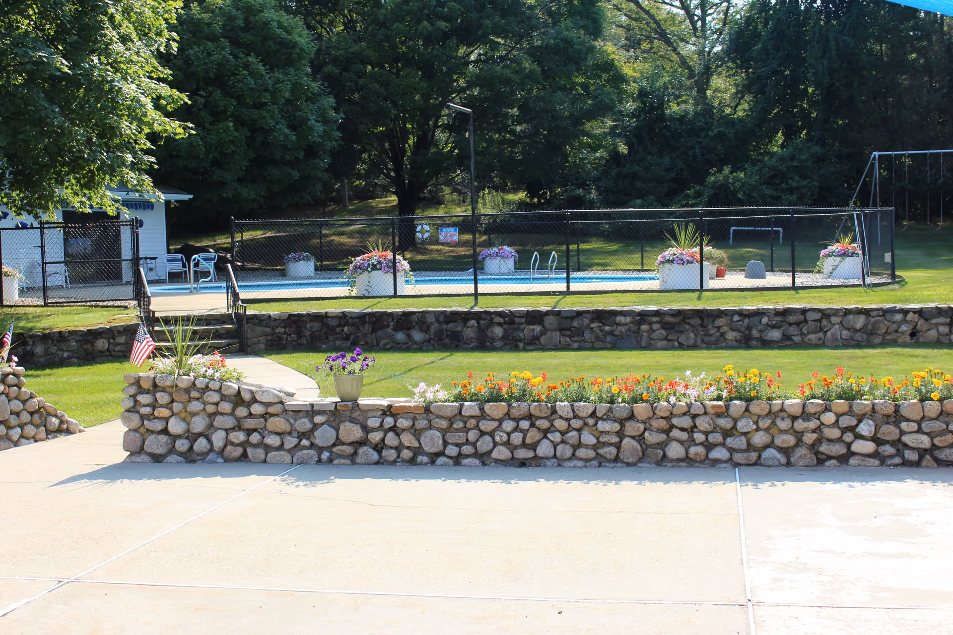 Stone-bordered flowerbeds and a grassy lawn in front of a fenced outdoor swimming pool with trees in the background.