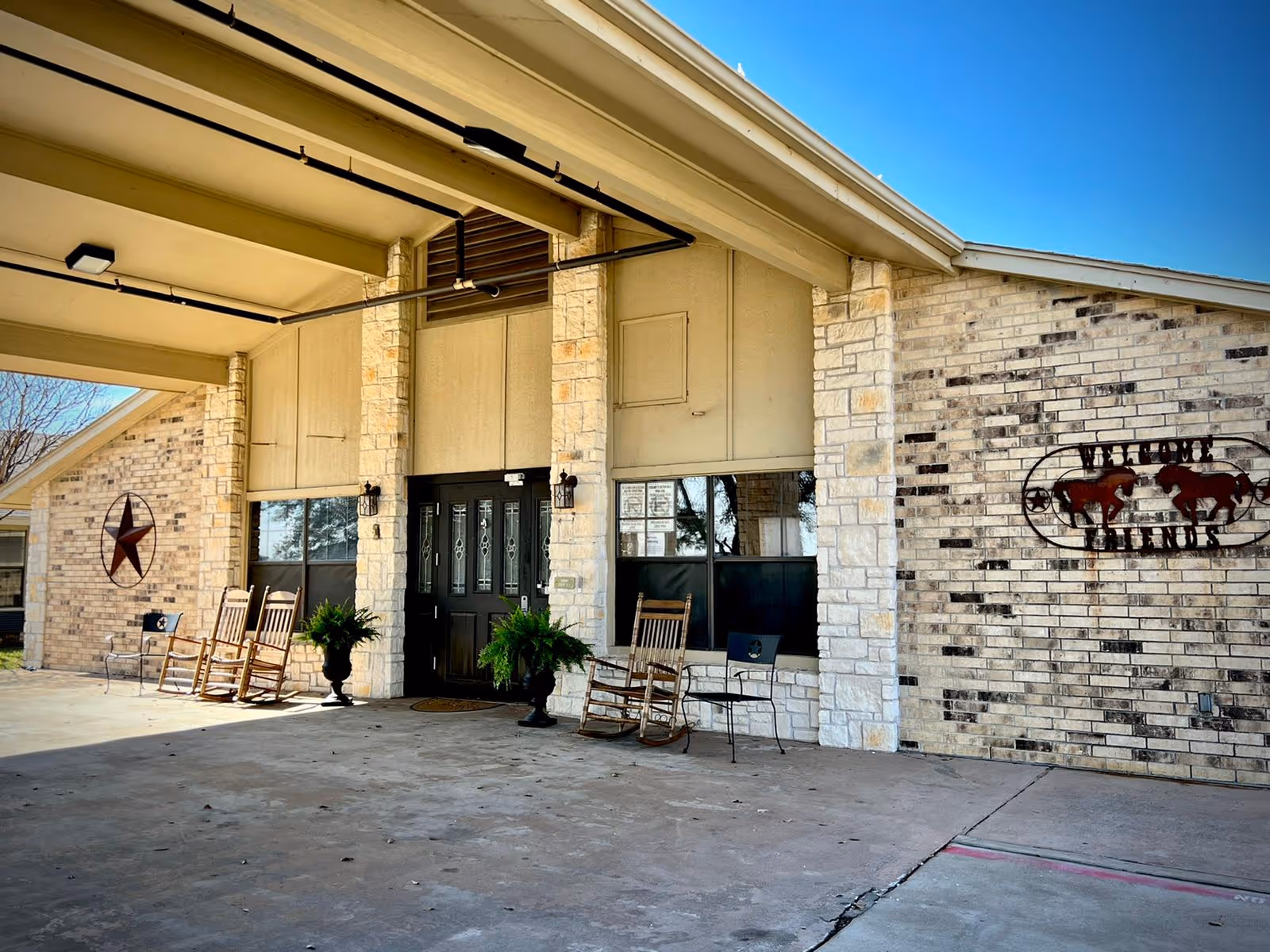 Front entrance of Cedar Ridge Rehabilitation and Healthcare Center featuring a covered driveway, stone and brick exterior walls, black double doors with glass panels, rocking chairs, metal chairs, potted plants, and decorative wall art including a large star and a sign with two horses and the words 'Wagon Wheel Ranch'.