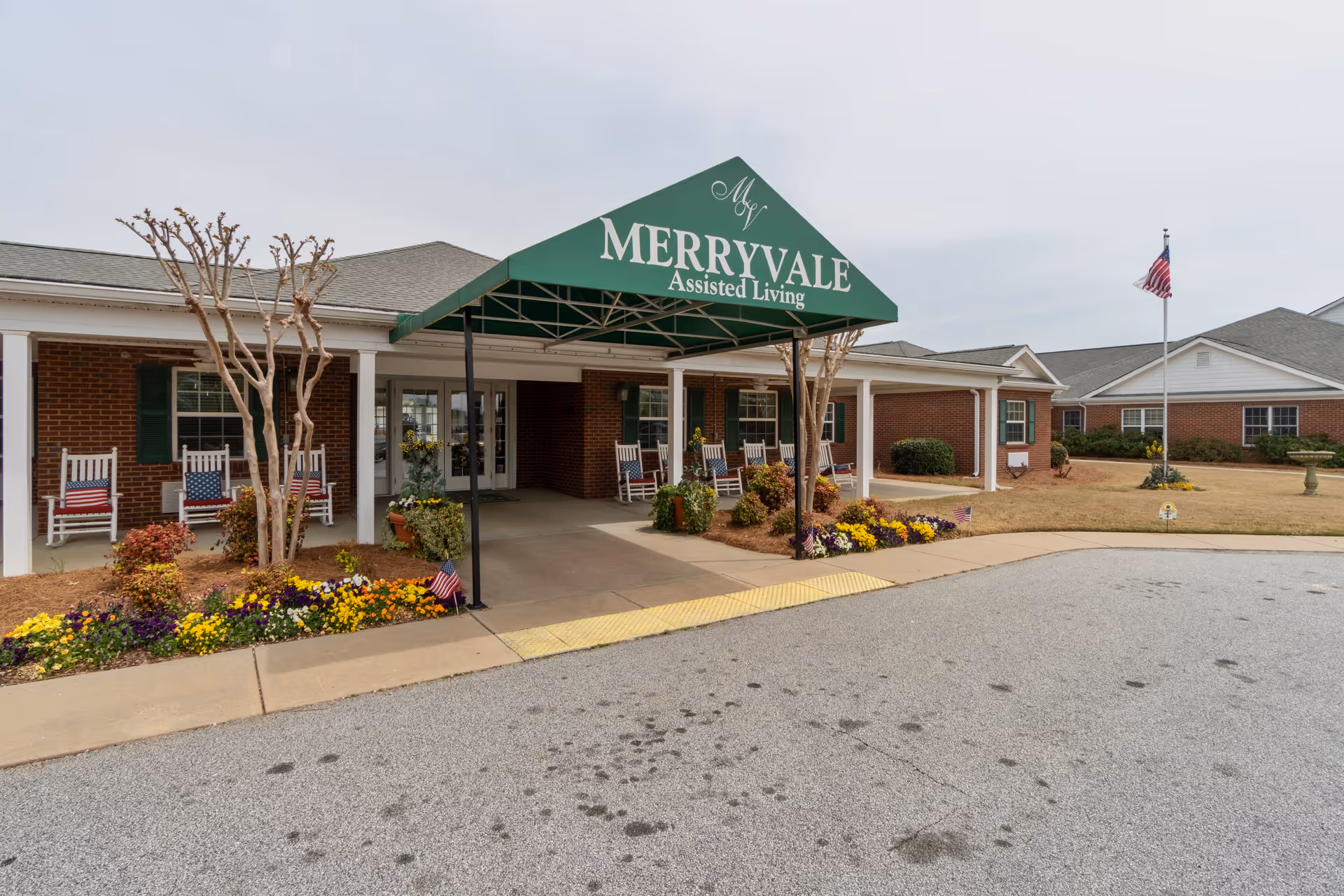 Exterior view of Merryvale Assisted Living facility showing a covered entrance with a green awning displaying the facility name. The building is brick with white columns and several rocking chairs with American flag cushions on the porch. There are flower beds with colorful flowers and a flagpole with an American flag on the lawn.