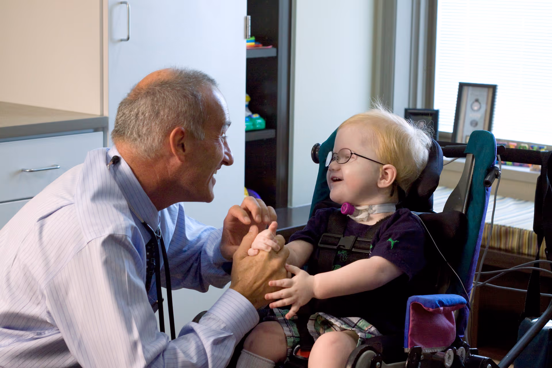 An elderly man wearing a light blue striped shirt and tie is smiling and holding hands with a young child in a wheelchair. The child is wearing glasses and a dark shirt, smiling back at the man. They are indoors in a room with cabinets and a window in the background.