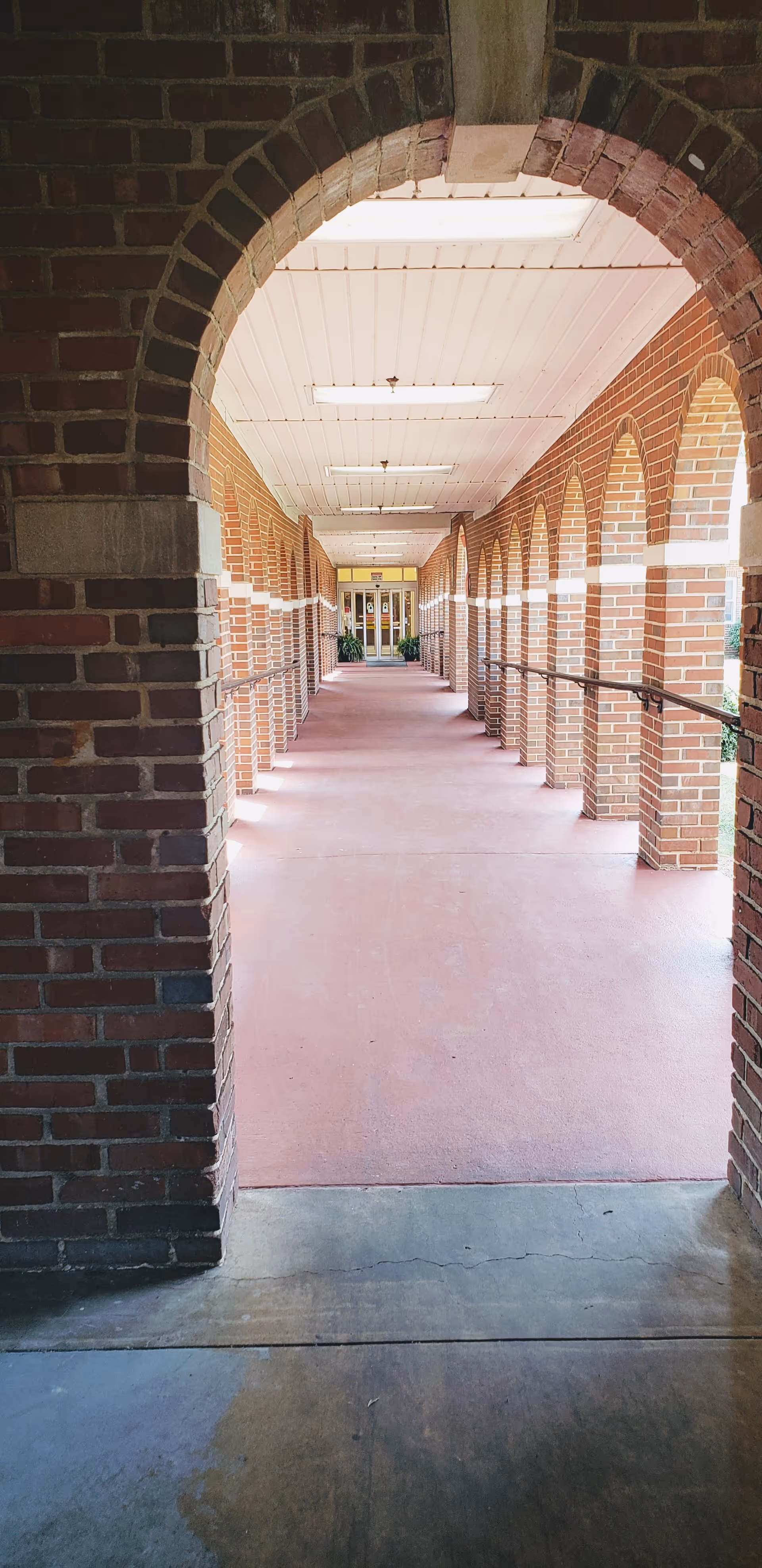 Covered brick arched walkway with handrails leading to glass entrance doors at the end.