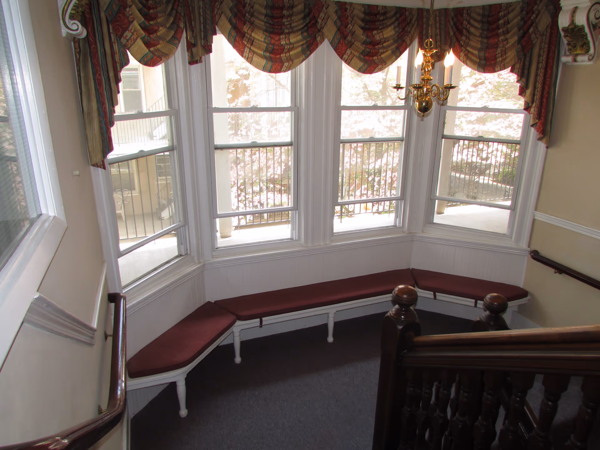 Interior corner seating area with a curved bench featuring maroon cushions beneath three large windows with patterned valance curtains. A brass chandelier hangs from the ceiling, and a wooden staircase railing is visible in the foreground.