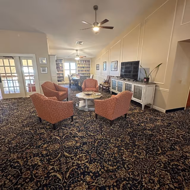 A cozy living room area with four patterned armchairs arranged around a round coffee table on a dark floral carpet. A flat-screen TV sits on a white cabinet against the wall, with a plant beside it. The room has beige walls with framed pictures and ceiling fans with lights. In the background, there is a dining area with a table and chairs near large windows and bookshelves.
