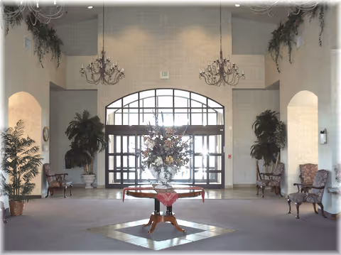 Spacious and elegant lobby area with a central round table adorned with a large floral arrangement. The room features high ceilings with two chandeliers, large glass entrance doors with an arched window above, and several potted plants and upholstered chairs arranged along the walls.