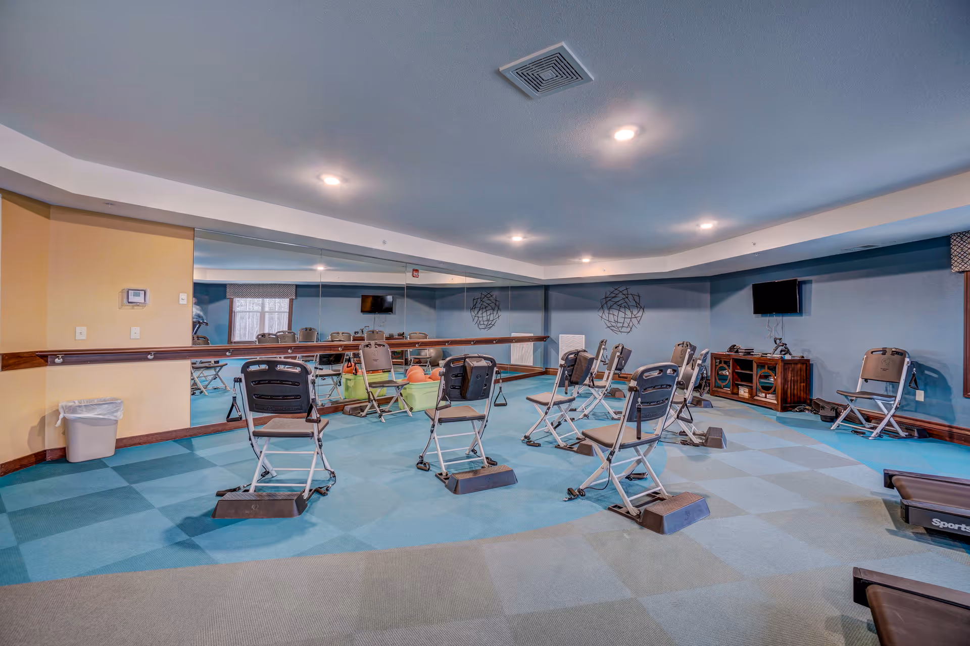 Exercise room with several folding chairs arranged in rows, each with resistance bands attached and footrests in front. The room has blue walls, a large mirror on one side, a TV mounted on the wall, and exercise equipment visible in the corner.