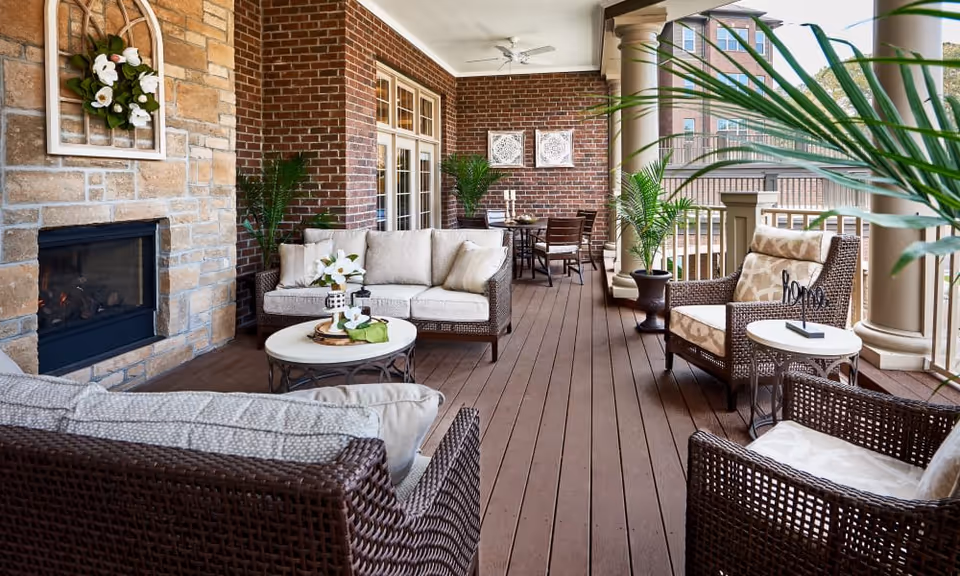 Covered outdoor patio area with wicker furniture including cushioned chairs and sofas, a round coffee table, potted plants, a stone fireplace with a floral decoration above it, and a dining table with chairs in the background. The patio has wooden flooring and is surrounded by brick walls and large columns.