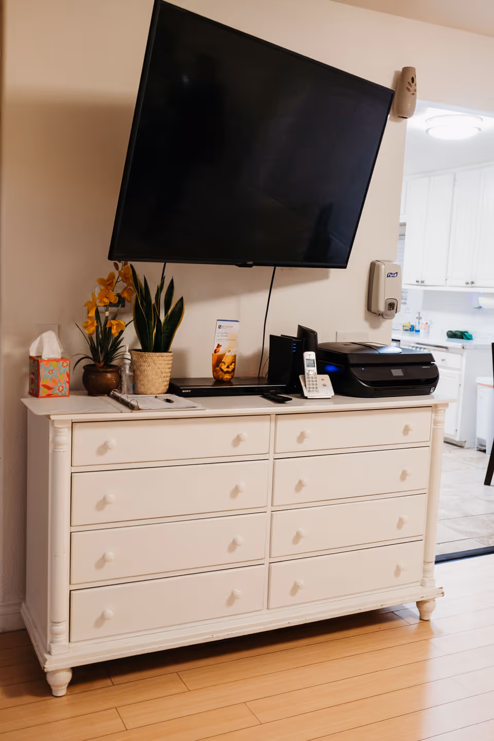 A white dresser with multiple drawers topped with a flat-screen TV mounted on the wall above it. On the dresser are a tissue box, two potted plants, a brochure, a cordless phone, a printer, and some electronic devices. The background shows a glimpse of a kitchen area with white cabinets and tiled floor.