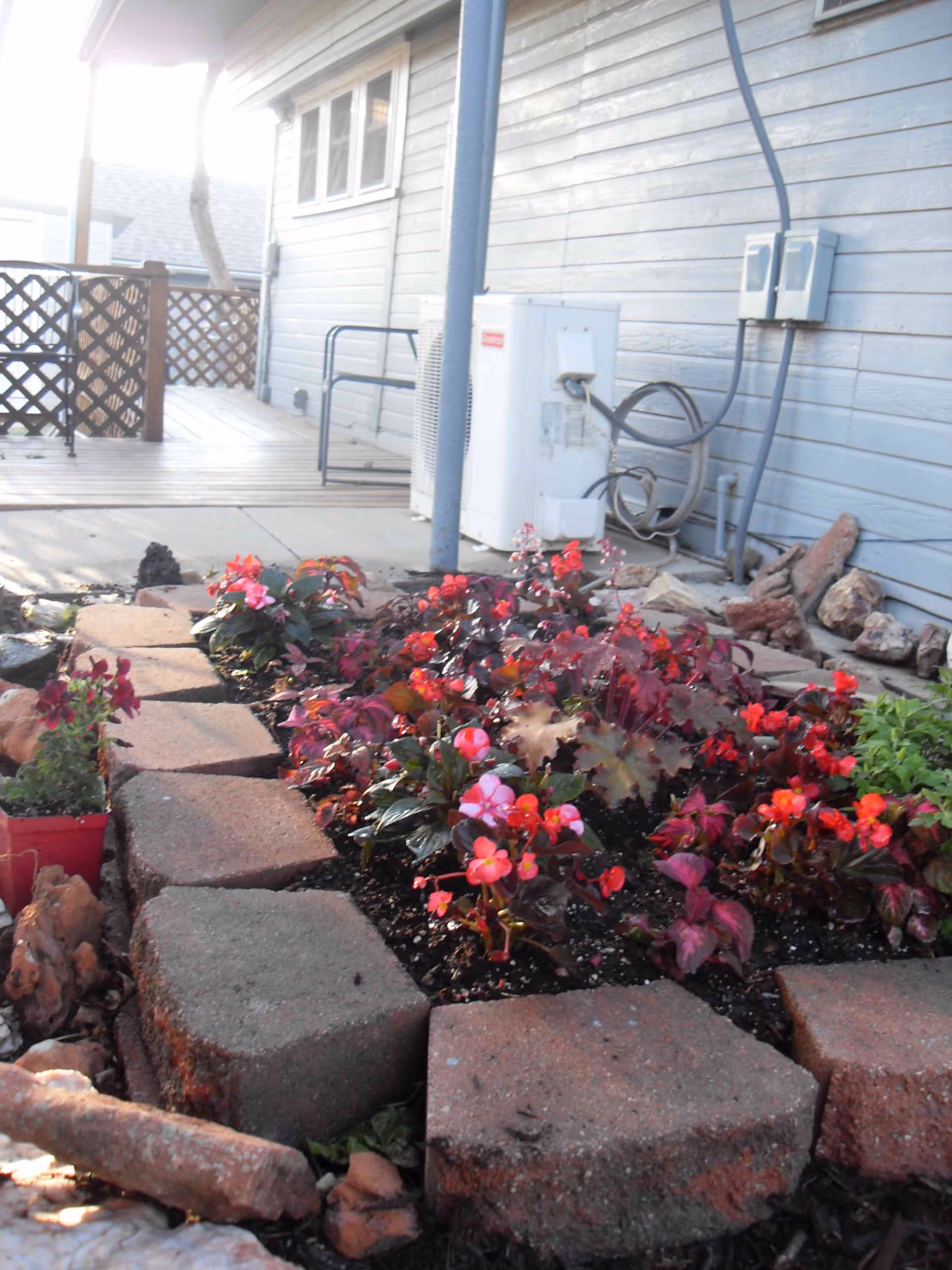 A small garden bed with red and pink flowers bordered by large rectangular stones, situated next to a light blue building exterior with visible utility meters and pipes. A wooden lattice fence and a concrete patio are also visible in the background.