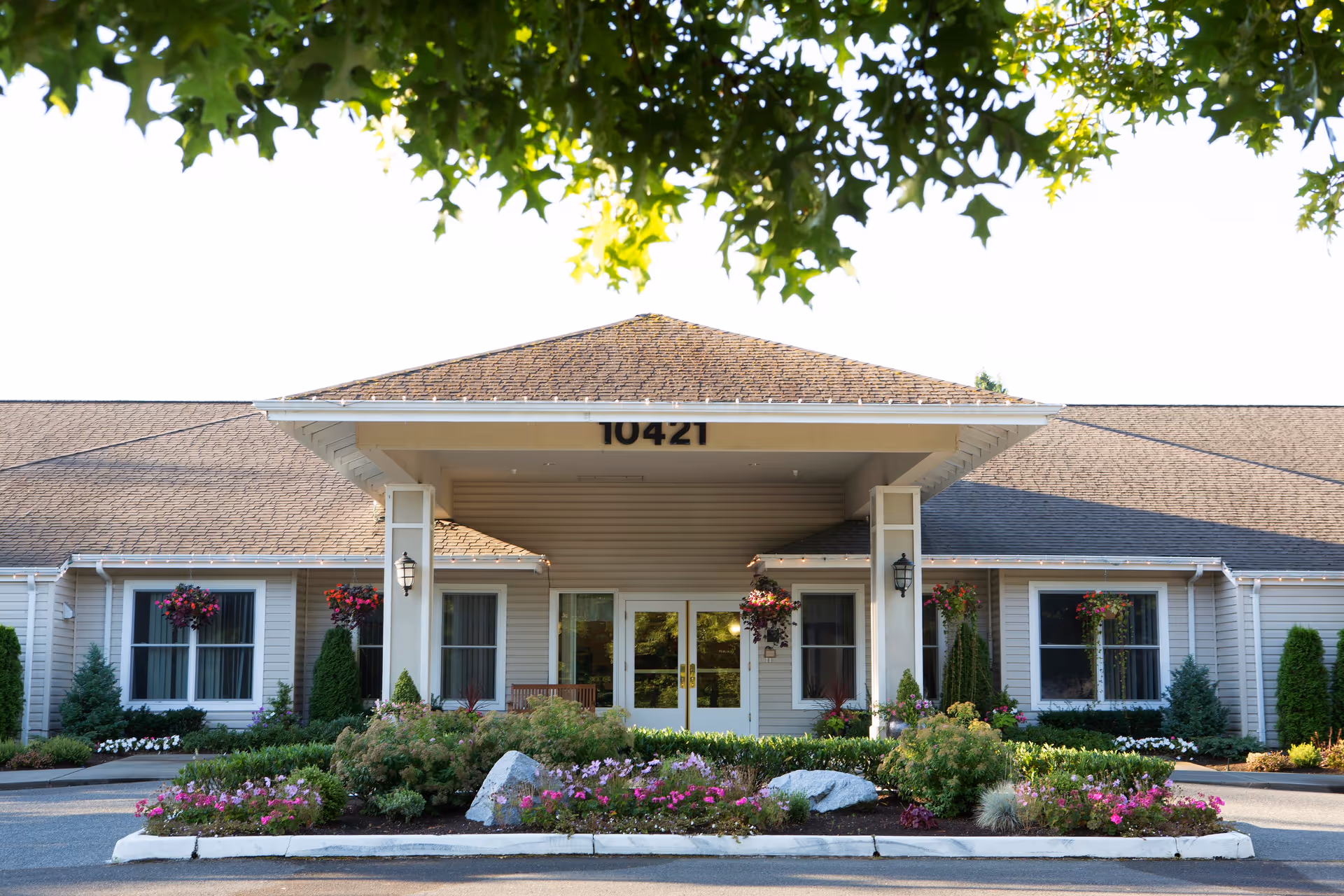 Front exterior view of a single-story building with a peaked roof and the number 10421 displayed above the entrance. The entrance is covered by a portico supported by two columns. There are windows on either side of the entrance with hanging flower baskets and well-maintained landscaping including bushes, flowers, and rocks in front of the building. Tree branches with green leaves frame the top of the image.