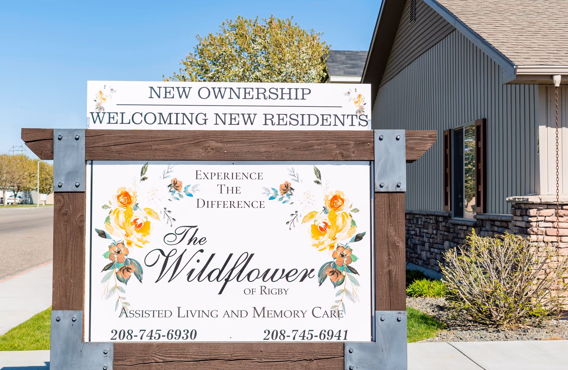 Outdoor wooden sign for The Wildflower of Rigby assisted living and memory care with floral designs in front of the facility building.