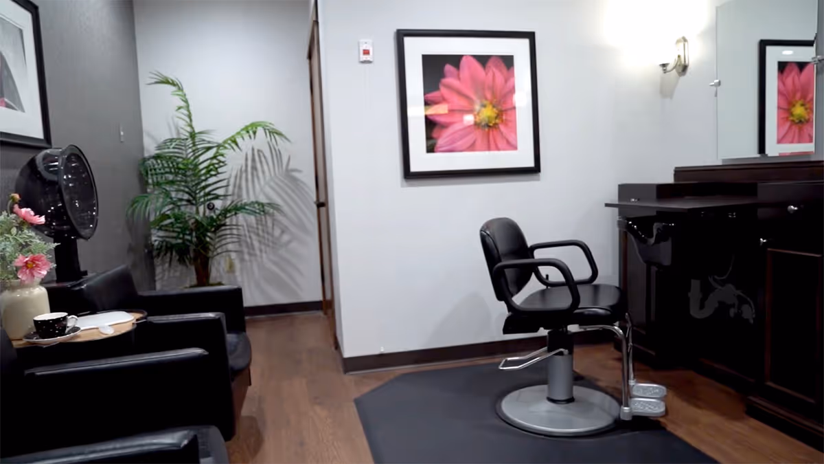 Interior view of a salon area in a senior living facility with a black salon chair in front of a dark wood vanity with a sink. There are two black leather chairs with a small table holding a cup and flowers, a hair dryer, a potted plant, and a framed picture of a pink flower on the wall.