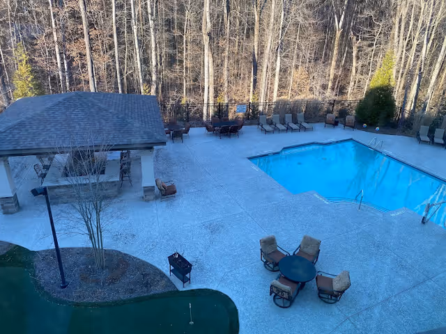 Outdoor pool area at Inspired Living Alpharetta featuring a rectangular swimming pool surrounded by lounge chairs and tables with chairs. There is a covered pavilion with bar stools and a small putting green nearby. Trees without leaves are visible in the background.