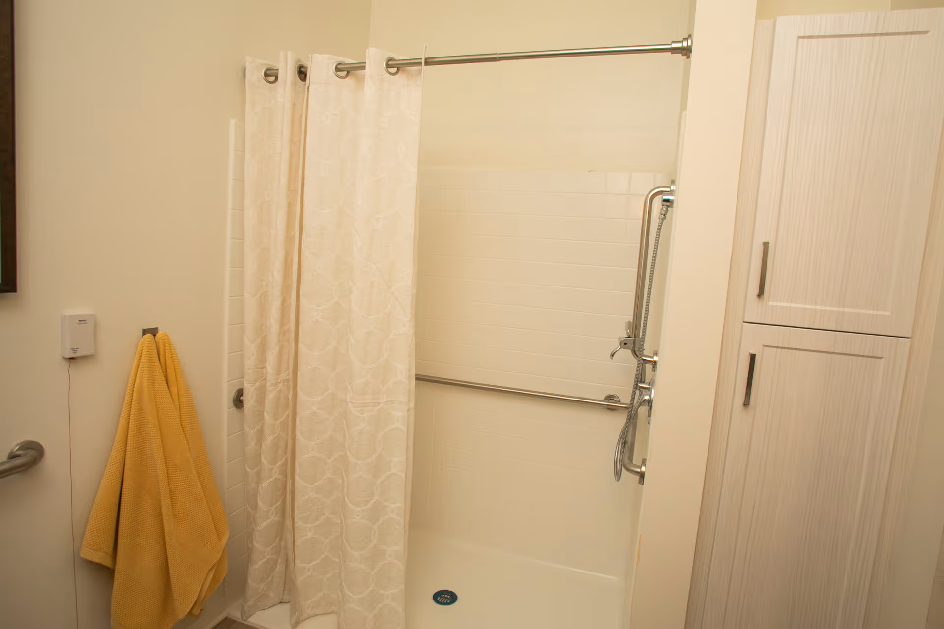 A bathroom shower area with a beige patterned shower curtain partially drawn. There is a metal grab bar inside the shower and a handheld showerhead mounted on the wall. A yellow towel hangs on a hook outside the shower, and a white cabinet with two doors is visible on the right side.