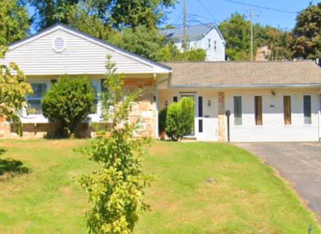 Single-story residential building with white siding and stone accents, surrounded by green grass and trees under a clear blue sky.