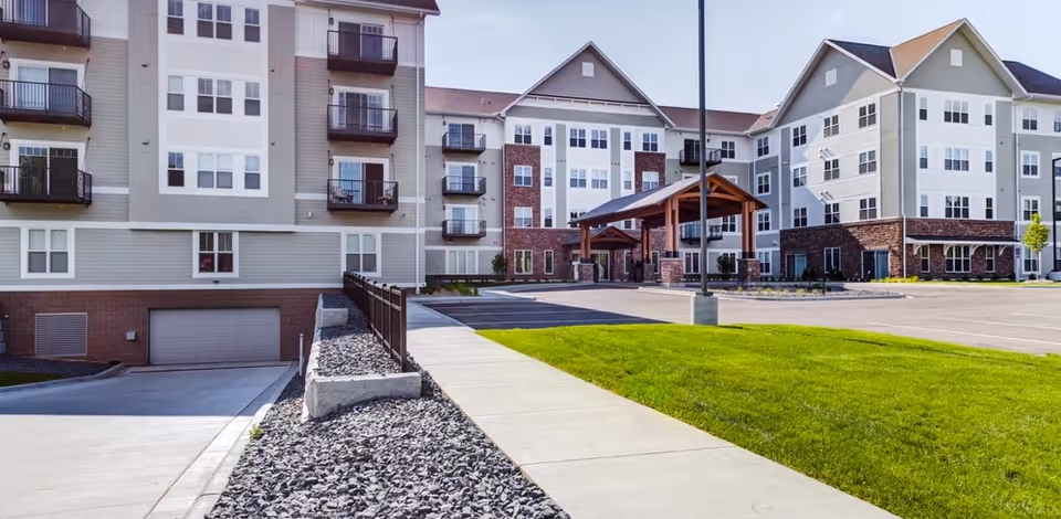 Front exterior of a multi-story senior living building with balconies, a covered entry porte-cochère, driveway and landscaped lawn.