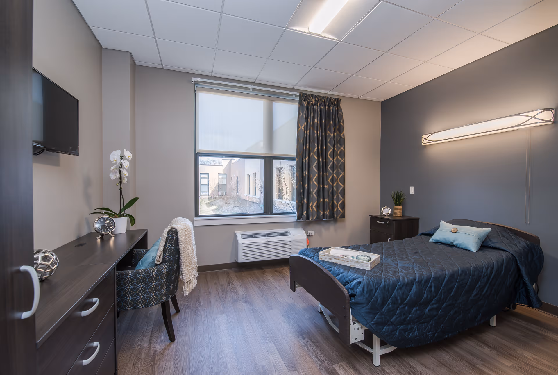 A neatly arranged bedroom in a senior living facility with a single bed covered in a dark blue quilt, a small bedside table with a plant and decorative item, a window with patterned curtains, a desk with a chair, a clock, and a potted orchid. The room has wood flooring and neutral-colored walls.