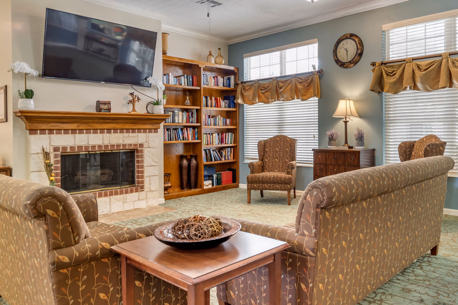 A cozy living room with patterned sofas and armchairs arranged around a wooden coffee table. There is a stone fireplace with a wooden mantle, above which a flat-screen TV is mounted. A wooden bookshelf filled with books is positioned next to the fireplace. The room has large windows with blinds and valances, a wall clock, and a table lamp on a wooden cabinet.