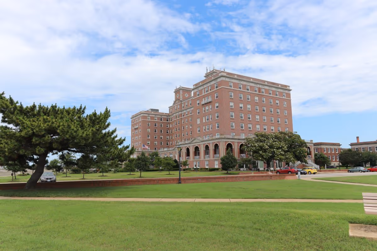 Large multi-story brick building with arched entrances set behind a grassy lawn and trees under a partly cloudy sky.