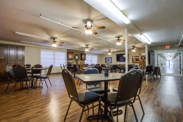 A spacious dining room with multiple tables and chairs arranged on a wooden floor. The room has ceiling fans with lights, large windows with blinds, and a hallway leading to other parts of the facility. There is a television mounted on the wall and cabinets along one side of the room.