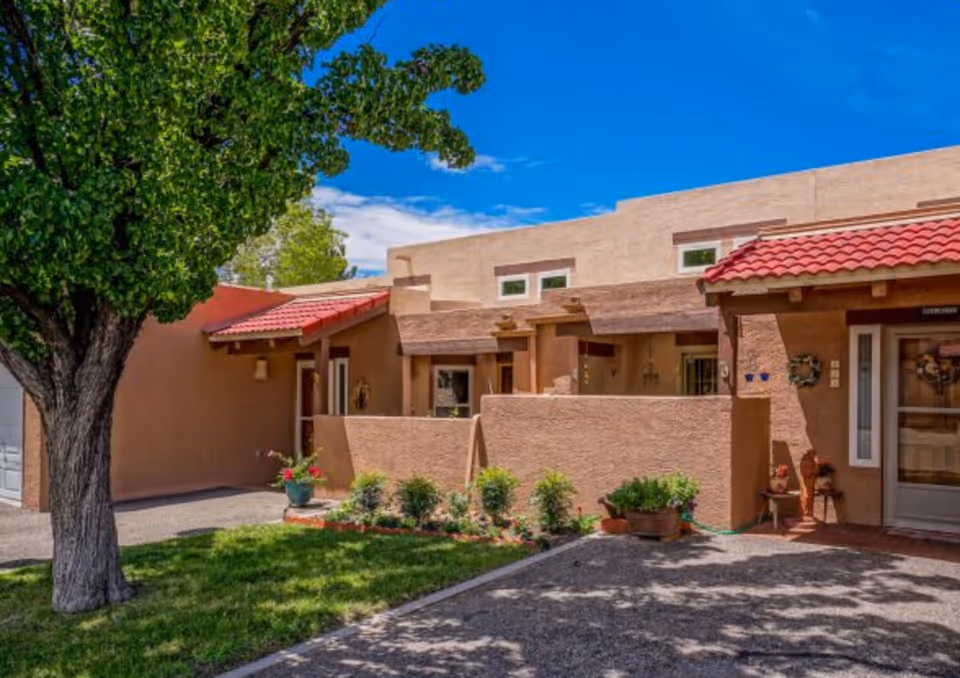 Single-story southwestern-style stucco building with red tile awnings, small front patios, potted plants and a large tree in the yard.