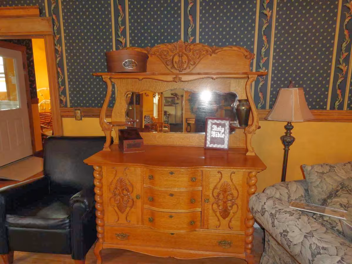 An interior room featuring a wooden dresser with ornate carvings and a large mirror. On top of the dresser are a small box, a framed Holy Bible, and a vase. To the left of the dresser is a black leather armchair, and to the right is a floral-patterned sofa with a floor lamp behind it. The walls have a two-tone design with yellow on the bottom and blue patterned wallpaper with floral vertical stripes on top.