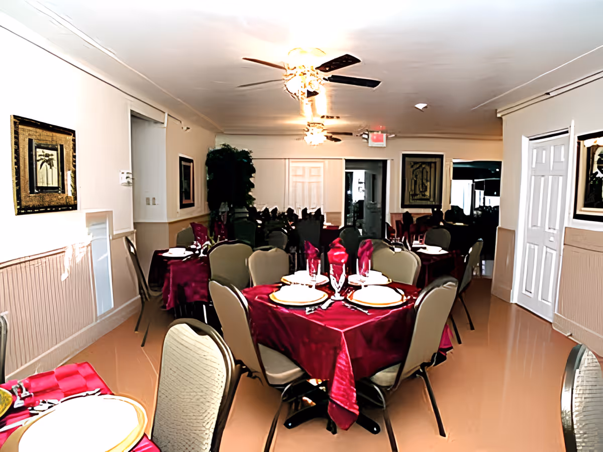 Interior view of a dining room with multiple tables covered in burgundy tablecloths, each set with plates, glasses, and folded napkins. The room has beige walls adorned with framed artwork, ceiling fans with lights, and beige cushioned chairs around the tables.