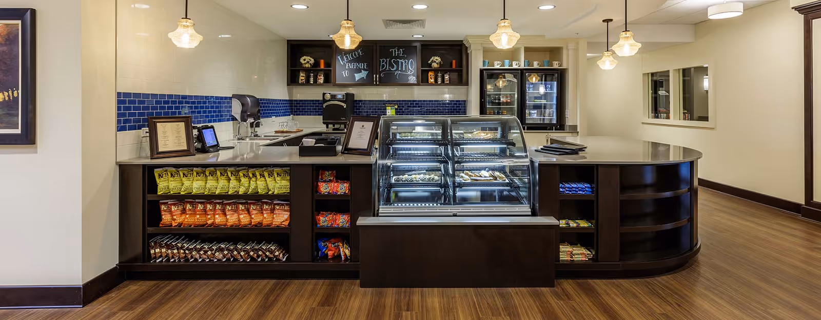 Interior view of a snack and beverage counter area named The Bistro with shelves stocked with chips and snacks, a glass display case with pastries, pendant lights hanging from the ceiling, and a blue tile backsplash behind the counter.