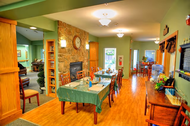 A cozy dining area in a senior living facility with a long table covered with a green tablecloth, set with placemats, cups, and utensils. The room features wooden chairs, a stone fireplace with a clock above it, green walls, and warm lighting. In the background, there is another dining table and a water dispenser on a wooden sideboard.