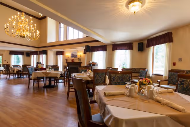 A spacious dining room with multiple tables covered in beige tablecloths, set with napkins, glasses, and small flower arrangements. The room features large windows with dark valances, wooden chairs with patterned upholstery, a chandelier, and a fireplace in the background.