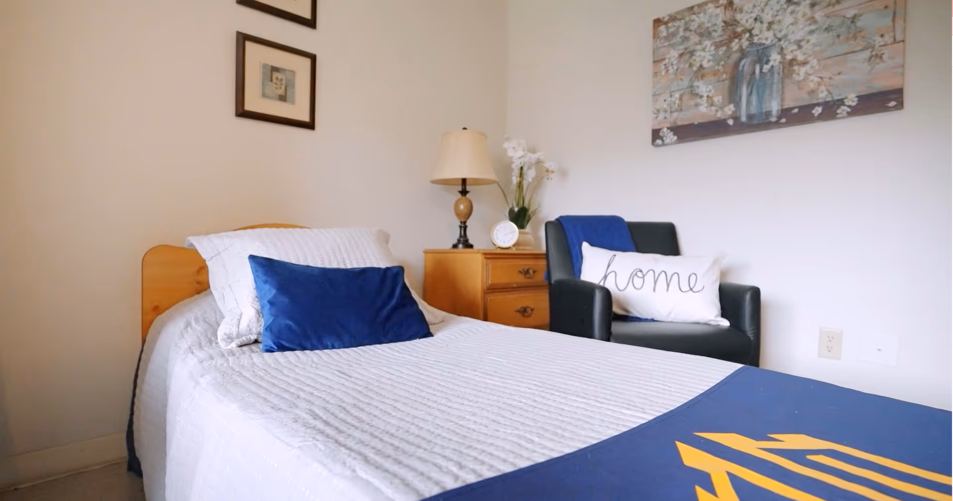 A neatly made single bed with white bedding and a blue pillow in a room. Next to the bed is a wooden nightstand with a lamp, a clock, and a vase with white flowers. A dark armchair with a white pillow that says 'home' and a blue throw blanket is positioned against the wall. Above the chair is a floral painting, and two framed pictures hang on the adjacent wall.