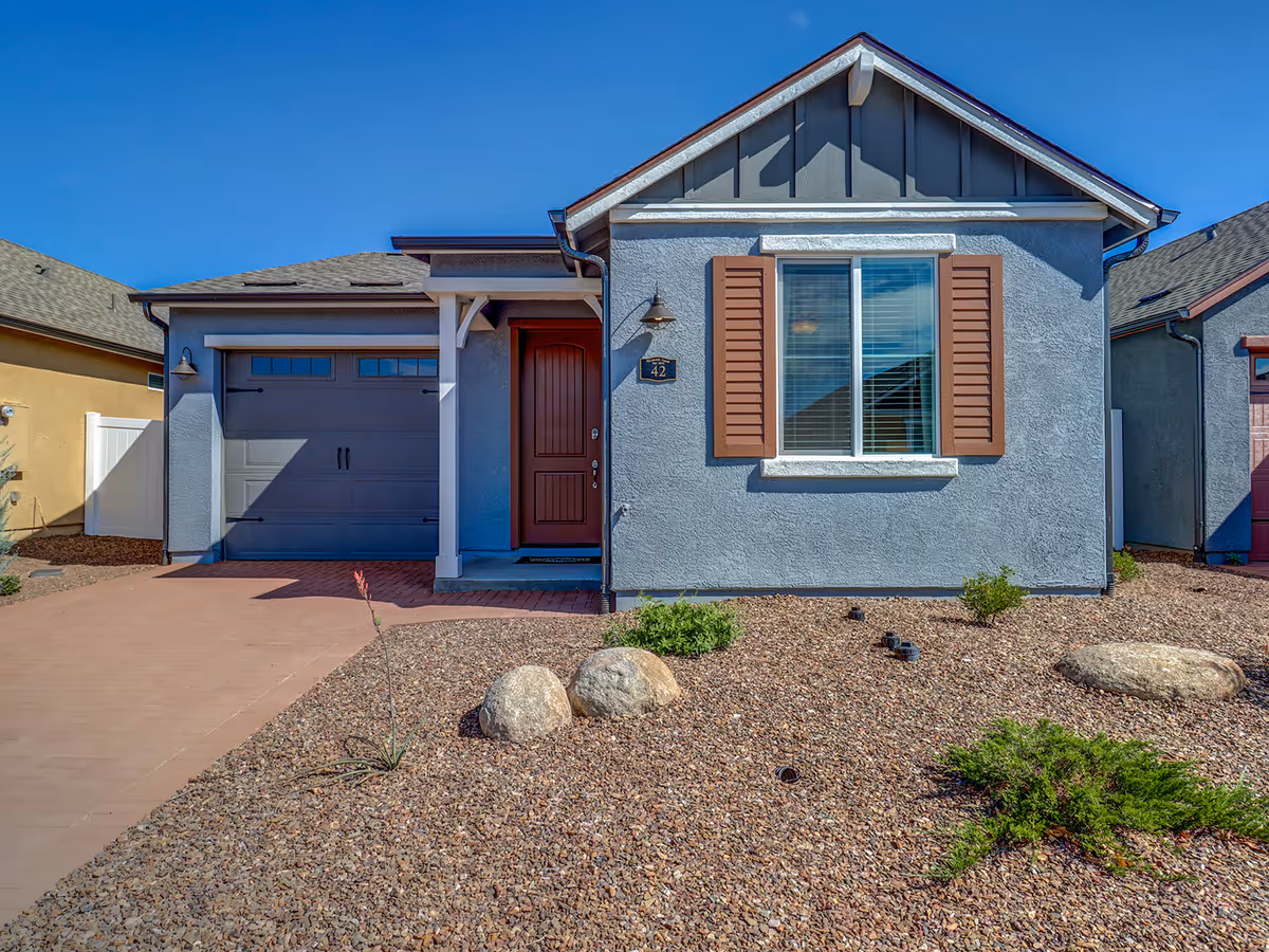 Front exterior of a single-story house with a garage, red front door, window with shutters, and rock landscaping.