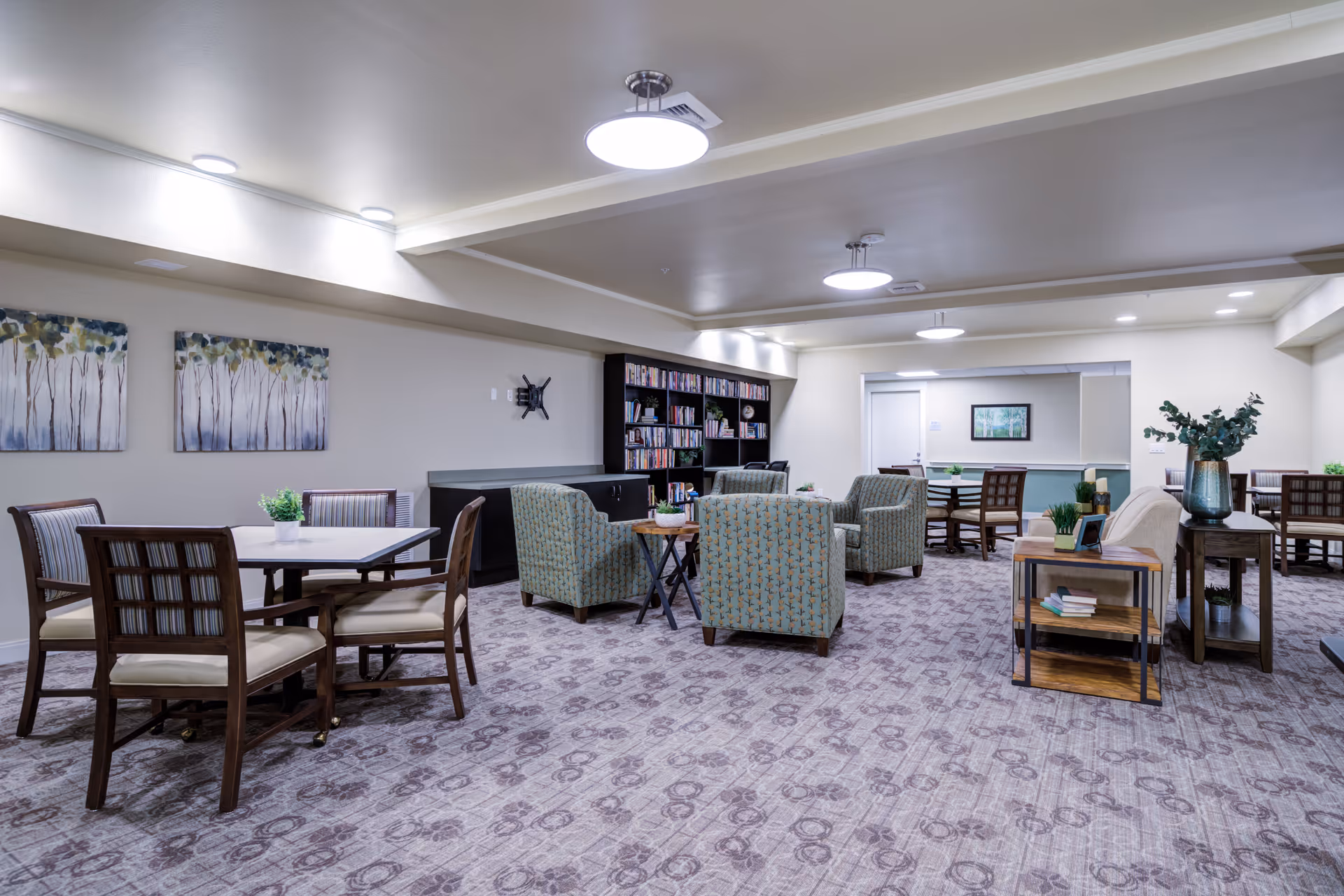 A spacious senior living community common area with multiple seating arrangements including tables with chairs and armchairs. The room features patterned carpet, soft lighting from ceiling fixtures, wall art depicting trees, and a bookshelf filled with books. There are also small tables with plants and decorative items.