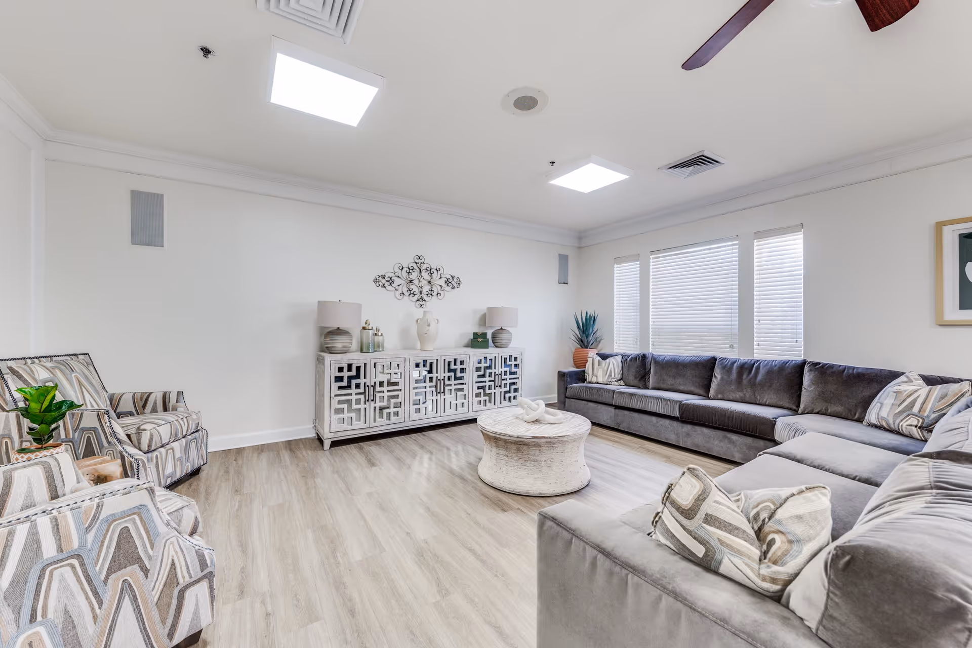 A bright and spacious living room with light wood flooring, a large gray sectional sofa with patterned pillows, two patterned armchairs, a round white coffee table, a decorative sideboard with lamps and vases, and three windows with blinds letting in natural light.