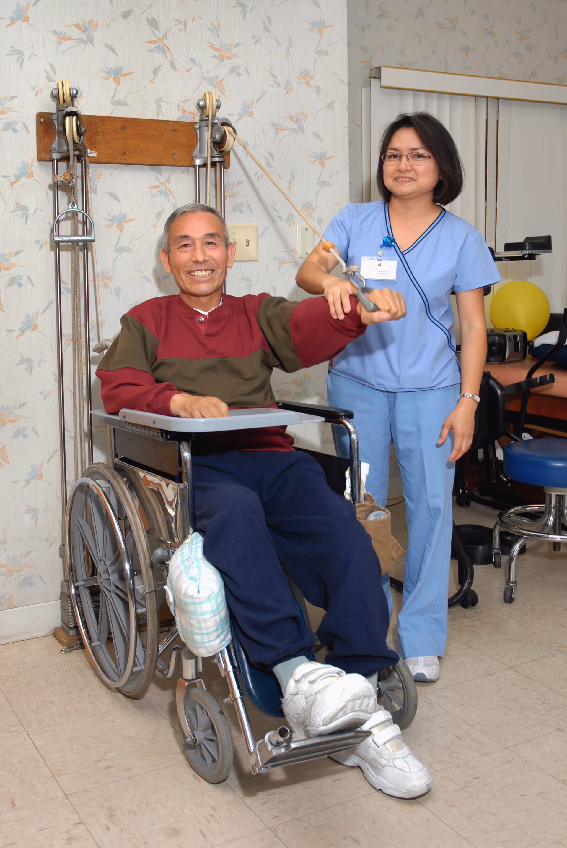 An elderly man in a wheelchair is smiling while performing a physical therapy exercise using a pulley system, assisted by a female healthcare worker in blue scrubs standing beside him in a rehabilitation room.