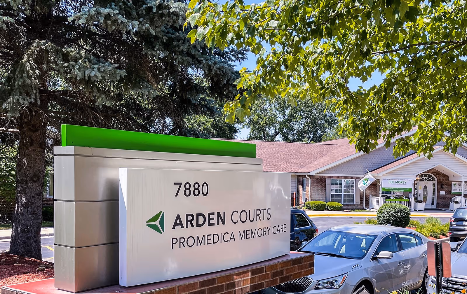 Outdoor view of Arden Courts - ProMedica Memory Care Community building with a large sign displaying the address 7880 and the facility name. Several cars are parked in front of the building, and trees with green leaves are visible around the area.