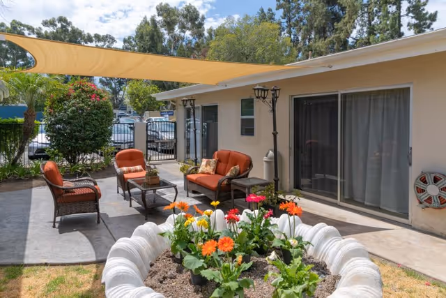 Outdoor patio area at Palomar Vista Healthcare Center with cushioned wicker chairs and a loveseat around a glass coffee table, a large planter with colorful flowers in the foreground, a beige shade sail overhead, and sliding glass doors leading into the building.