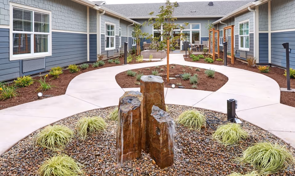 Outdoor courtyard area at Battle Creek Memory Care featuring a stone water fountain surrounded by small plants and gravel. The courtyard is enclosed by a building with blue siding and white-trimmed windows. There are paved walkways, landscaped garden beds, and outdoor seating visible in the background.