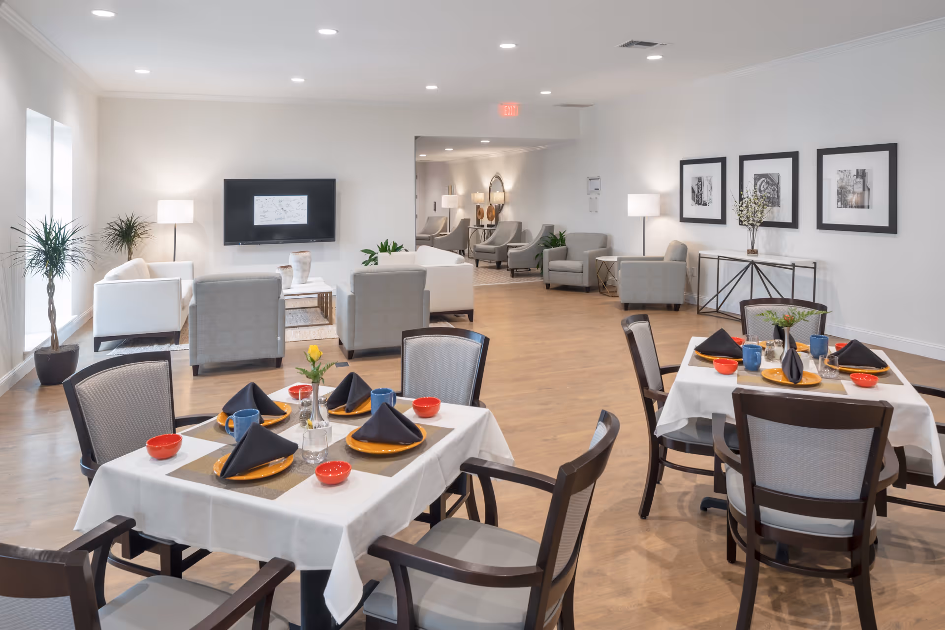 A bright and spacious senior living facility common area featuring two dining tables set with orange plates, black napkins, blue mugs, and red bowls. The room has light wood flooring, white walls, and modern decor including framed black and white photos on the wall. In the background, there is a lounge area with white and gray armchairs, a wall-mounted TV, floor lamps, and potted plants.