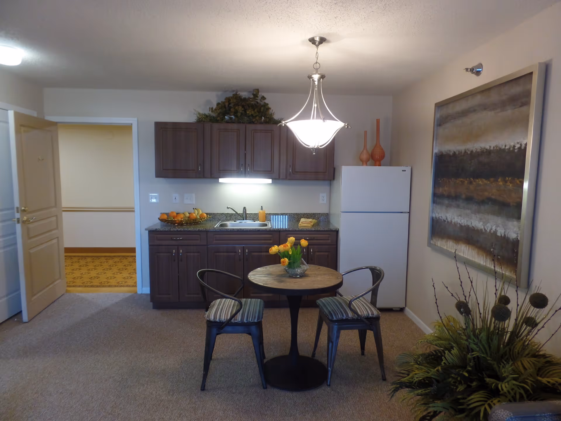 A small kitchen area with dark wooden cabinets, a white refrigerator, and a granite countertop with a sink. A round wooden table with two metal chairs with striped cushions is in front of the kitchen. A light fixture hangs from the ceiling above the table. There is a large abstract painting on the wall to the right and a plant in the corner.