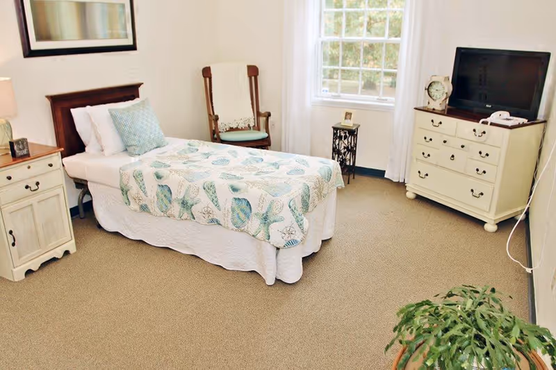 Bright single-occupancy bedroom with a twin bed, nightstand, rocking chair, dresser with TV, and a window.