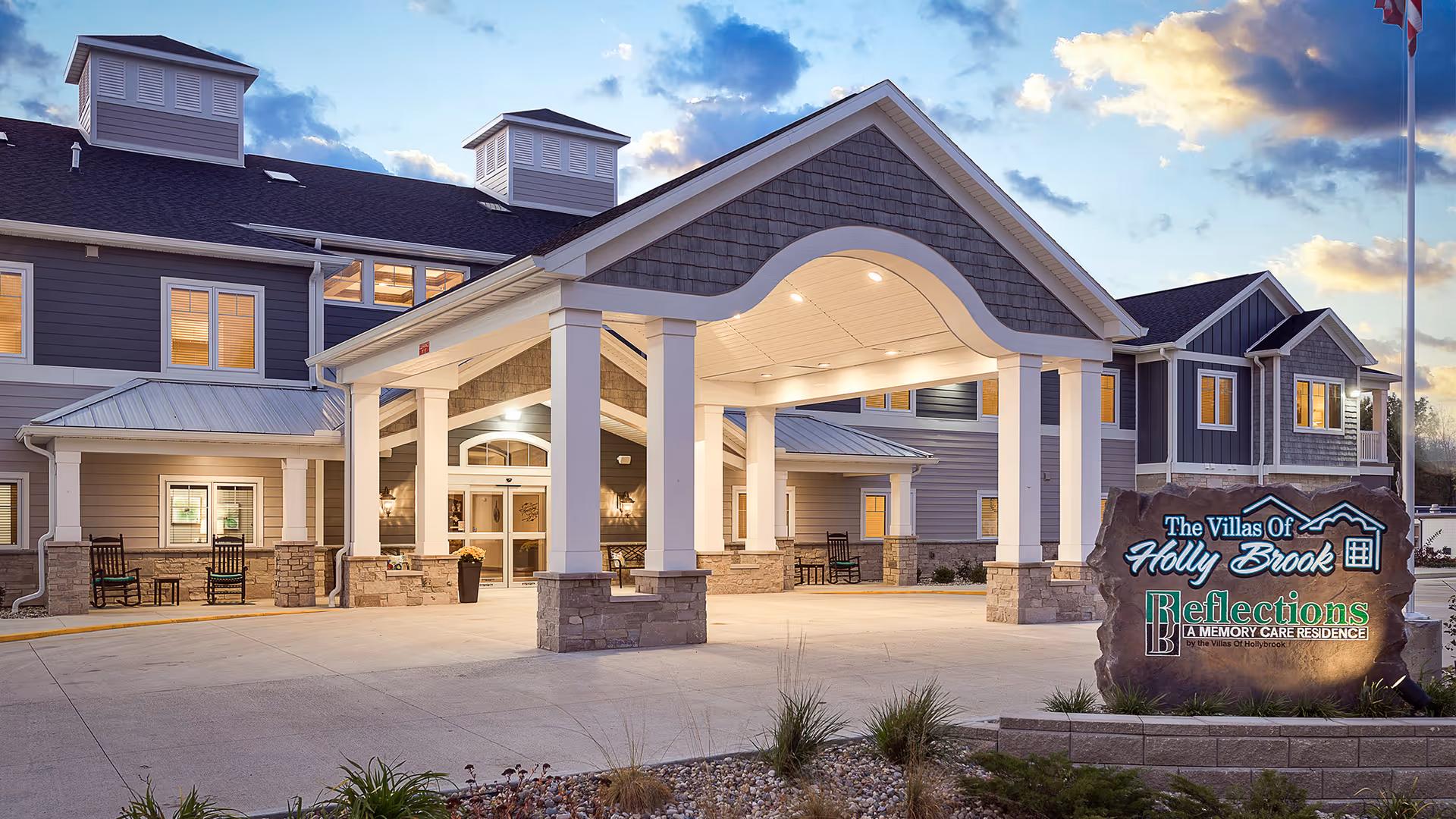 Exterior view of Villas of Holly Brook Assisted Living & Memory Care facility in Collinsville, IL during dusk. The building features a covered entrance with white pillars and stone accents. There are rocking chairs on the porch and a large stone sign displaying the facility's name and memory care residence information. The sky is partly cloudy with a warm glow from the setting sun.